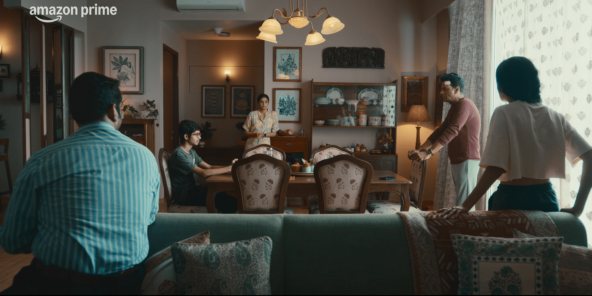 A tense family moment unfolds in a warmly lit living room, with several people standing and sitting around a dining table, appearing to argue or discuss something serious.