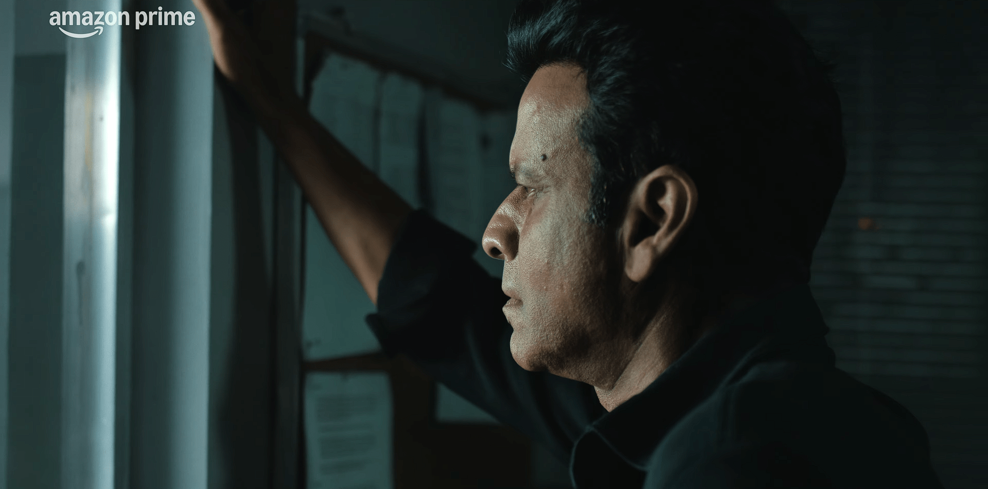 Shrikant stands in dim light, staring out of a window with a troubled and reflective expression.