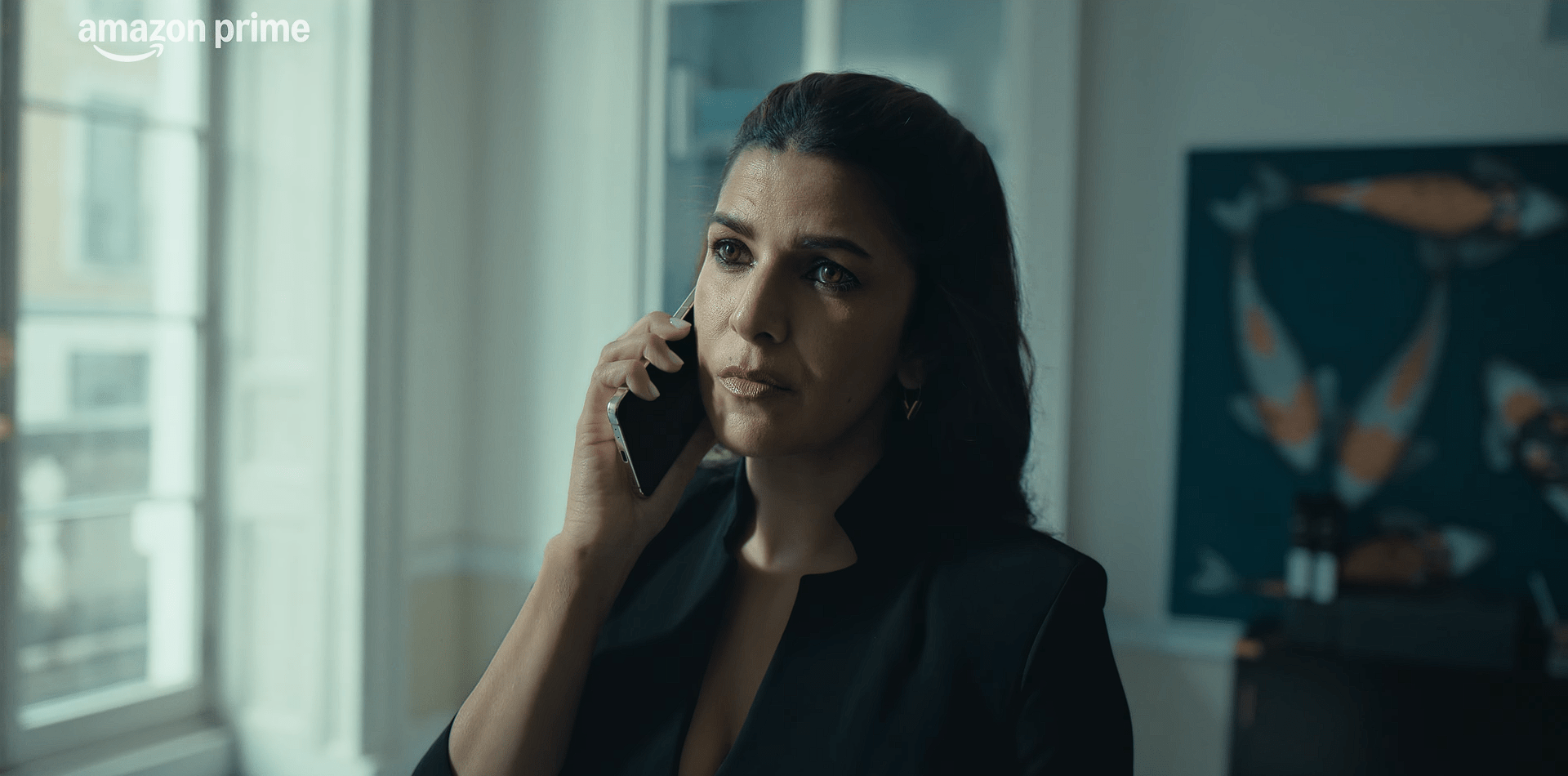 A woman dressed in formal clothing holds a phone to her ear, listening intently with a concerned, focused expression in a softly lit room.