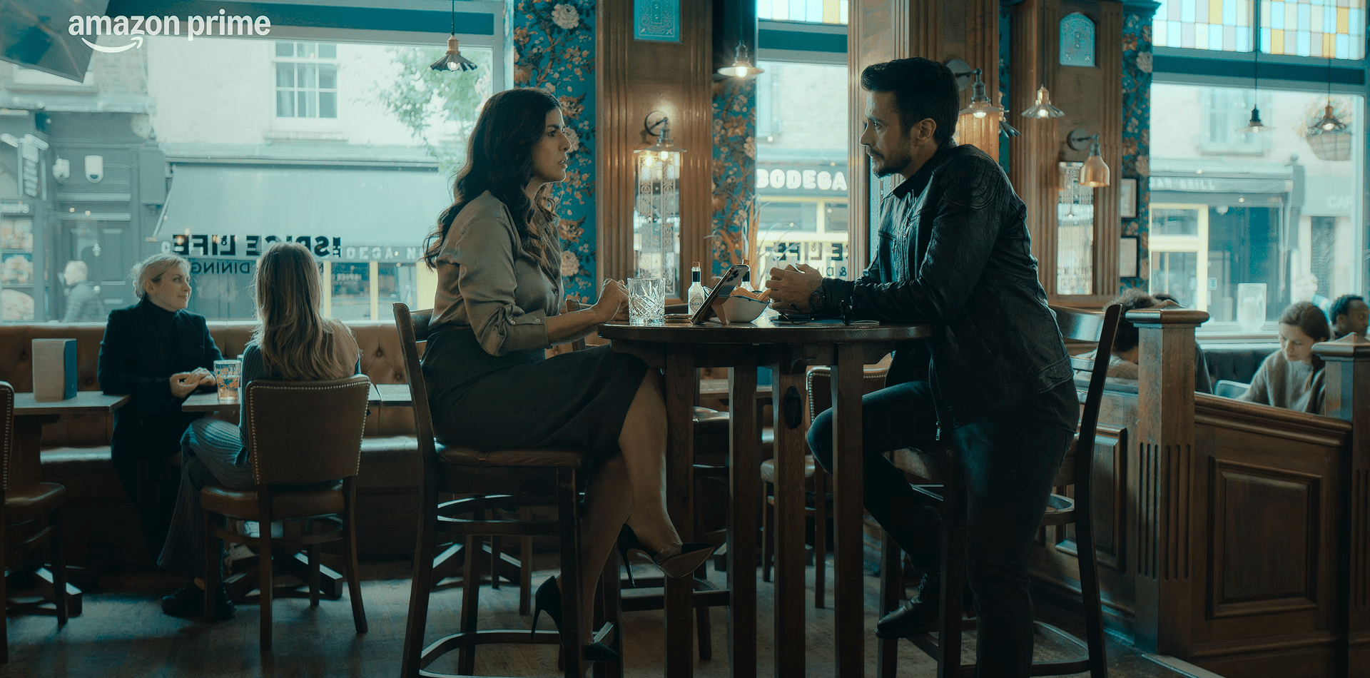 A man and a woman sit across from each other at a café table, engaged in a serious conversation, with other customers visible in the background.