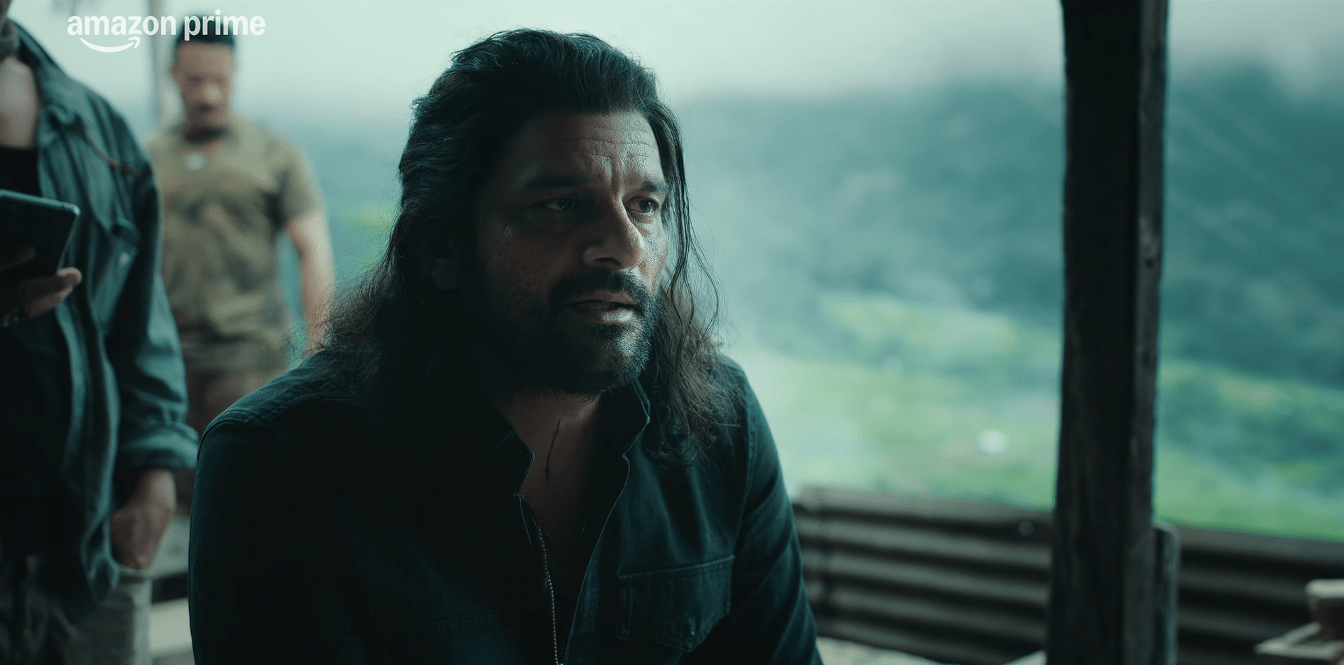 A rugged-looking man with long hair speaks with a tense expression, set against a misty hillside landscape.