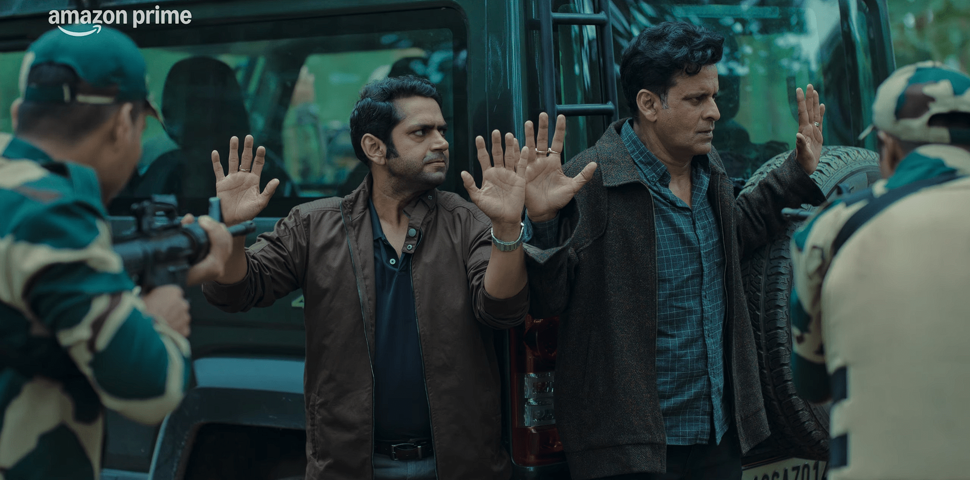 Two men stand with their hands raised beside a vehicle as armed soldiers aim weapons at them in a forested area.