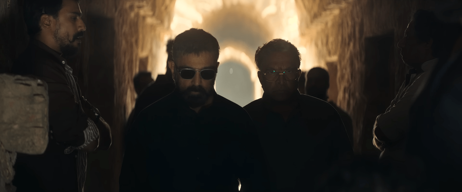 Sakthi wearing sunglasses walks through a dim stone corridor with several men around him, highlighted by warm light in the distance.