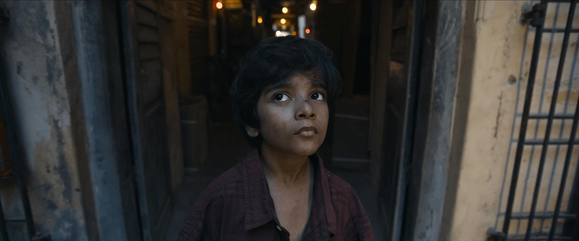 Young Amaran with messy hair and a bruised nose stands in a narrow alleyway, looking upward with a mix of fear and curiosity.