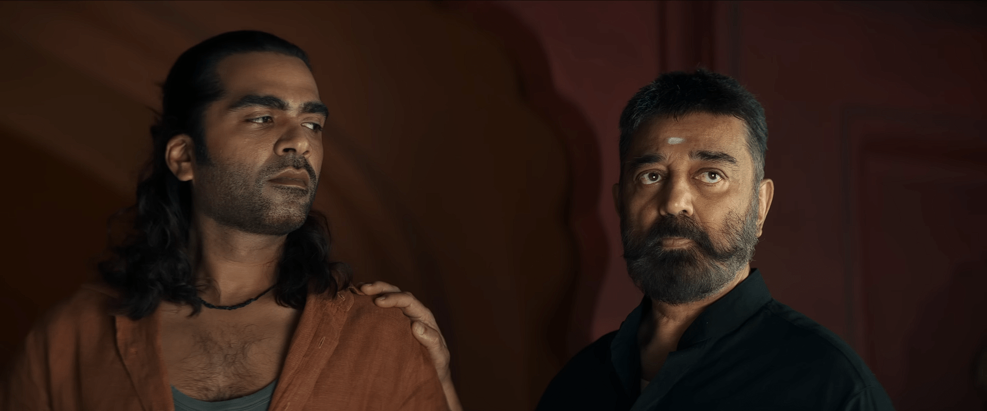Adult Amaran with long hair stands beside Sakthivel dressed in black, both looking serious as they face forward against a dark background.