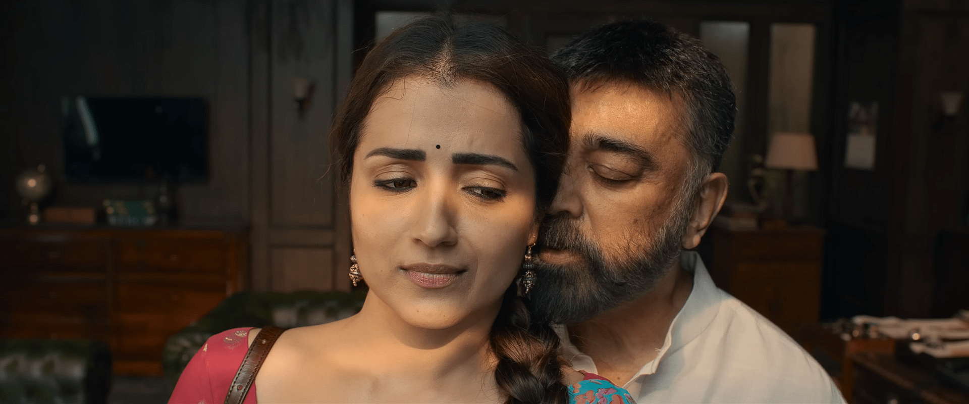 Indhrani looks down with a worried expression while Sakthivel stands close behind her in a warmly lit room filled with wooden furniture.