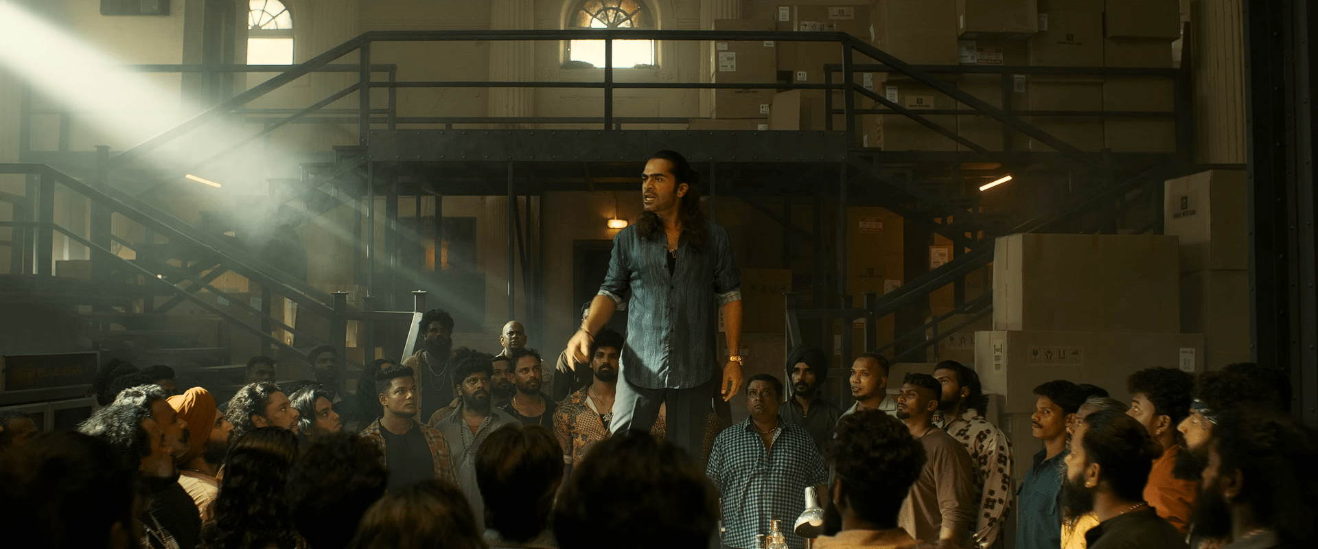 Amaran stands on a platform addressing a large group of men inside a dimly lit warehouse, with sunlight cutting through the dusty air.