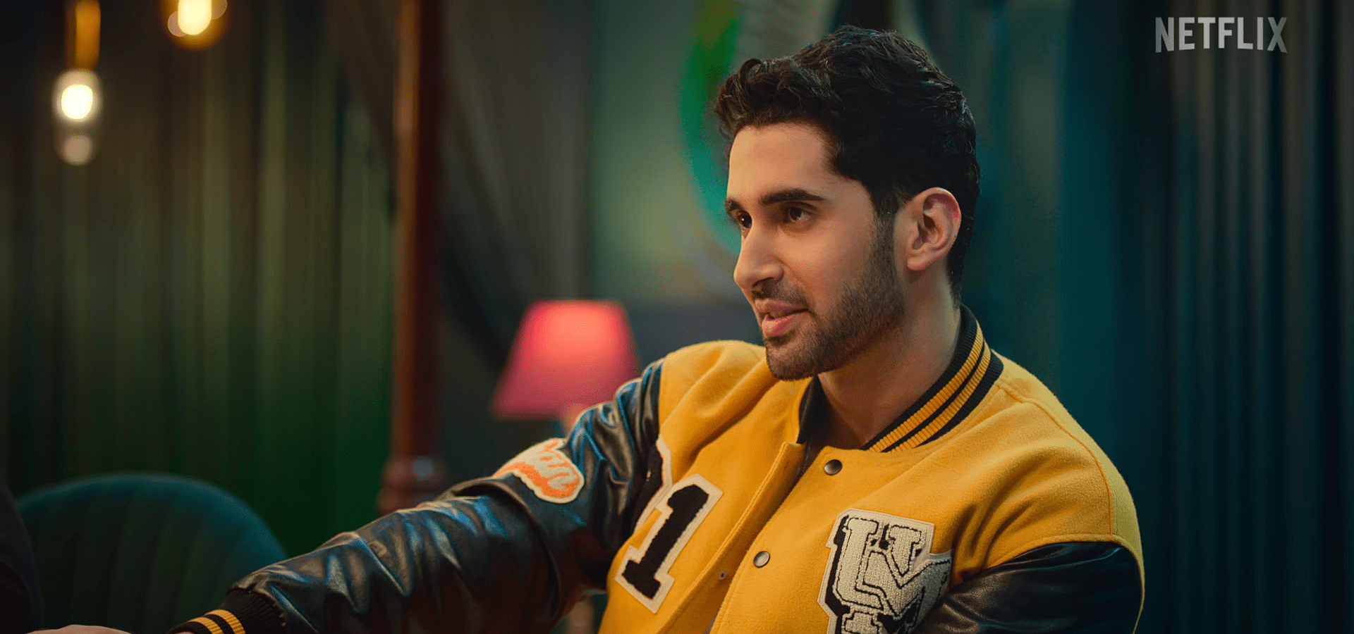 Lakshya Lalwani in a yellow varsity jacket speaks during a discussion, seated in a warmly lit indoor setting.