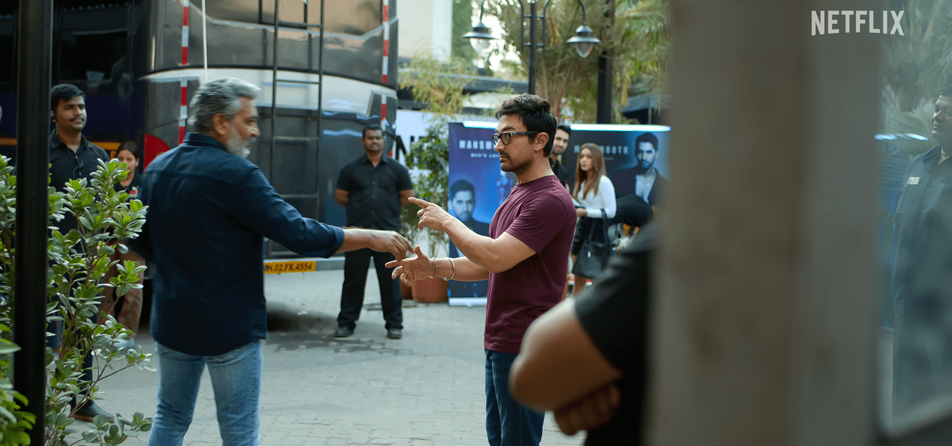 S. S. Rajamouli and Amir Khan having a brief interaction outdoors near a vanity van area, with security personnel and film-set posters around them.