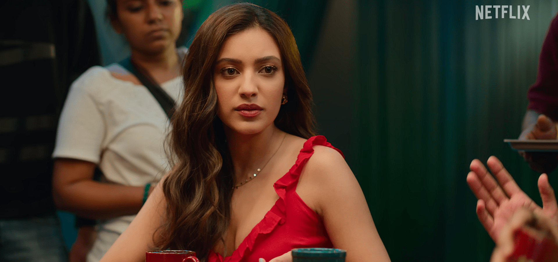 Sahher Bambba in a red sleeveless top listens attentively during a conversation, seated at a table with crew members visible behind her.