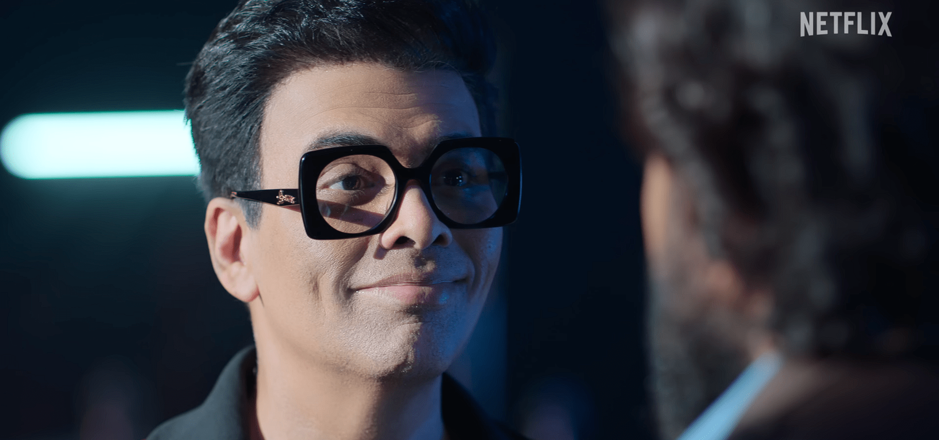 Karan Johar wearing thick square glasses smiles while standing under cool blue lighting, facing someone in close conversation.