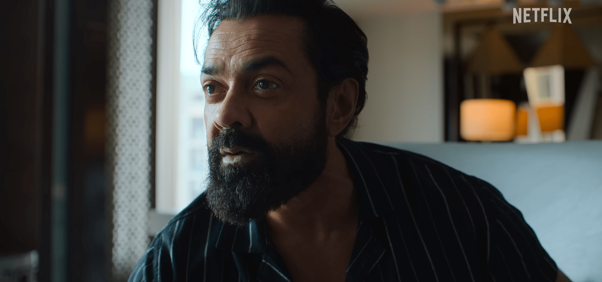 Bobby Deol leans forward while speaking, indoors near a window with soft daylight illuminating his face.