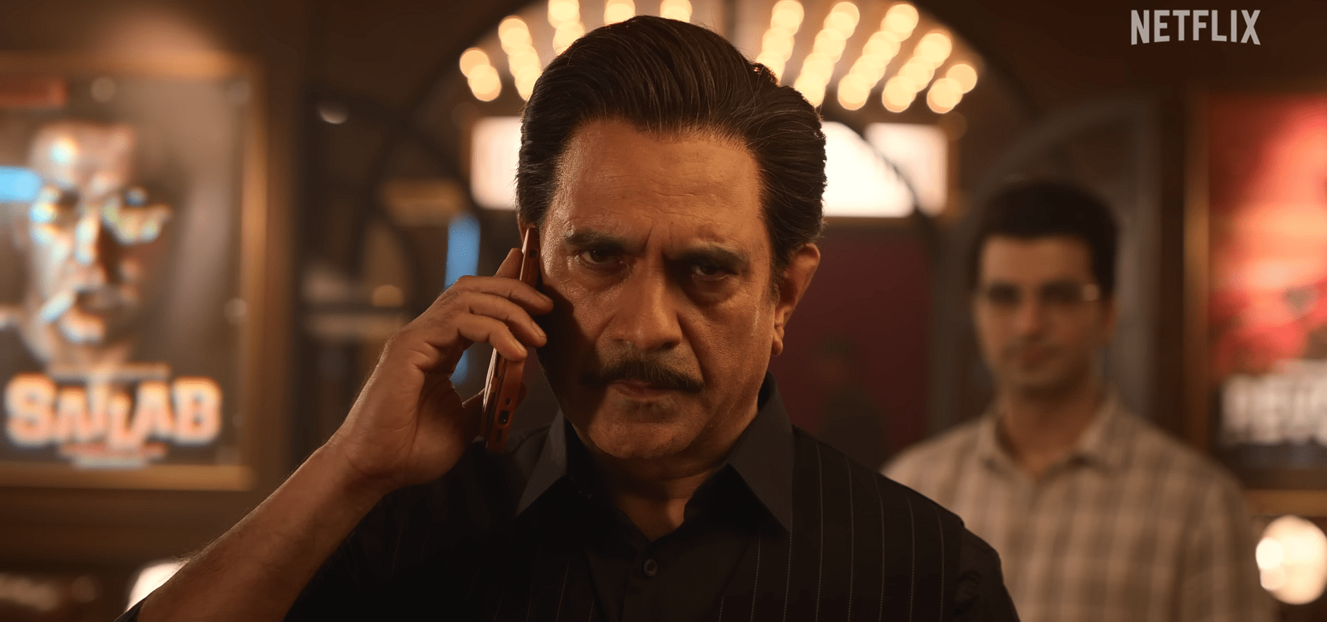 Manish Chaudhari in a dark vest talks on the phone inside a dim theatre-like room with movie posters in the background.