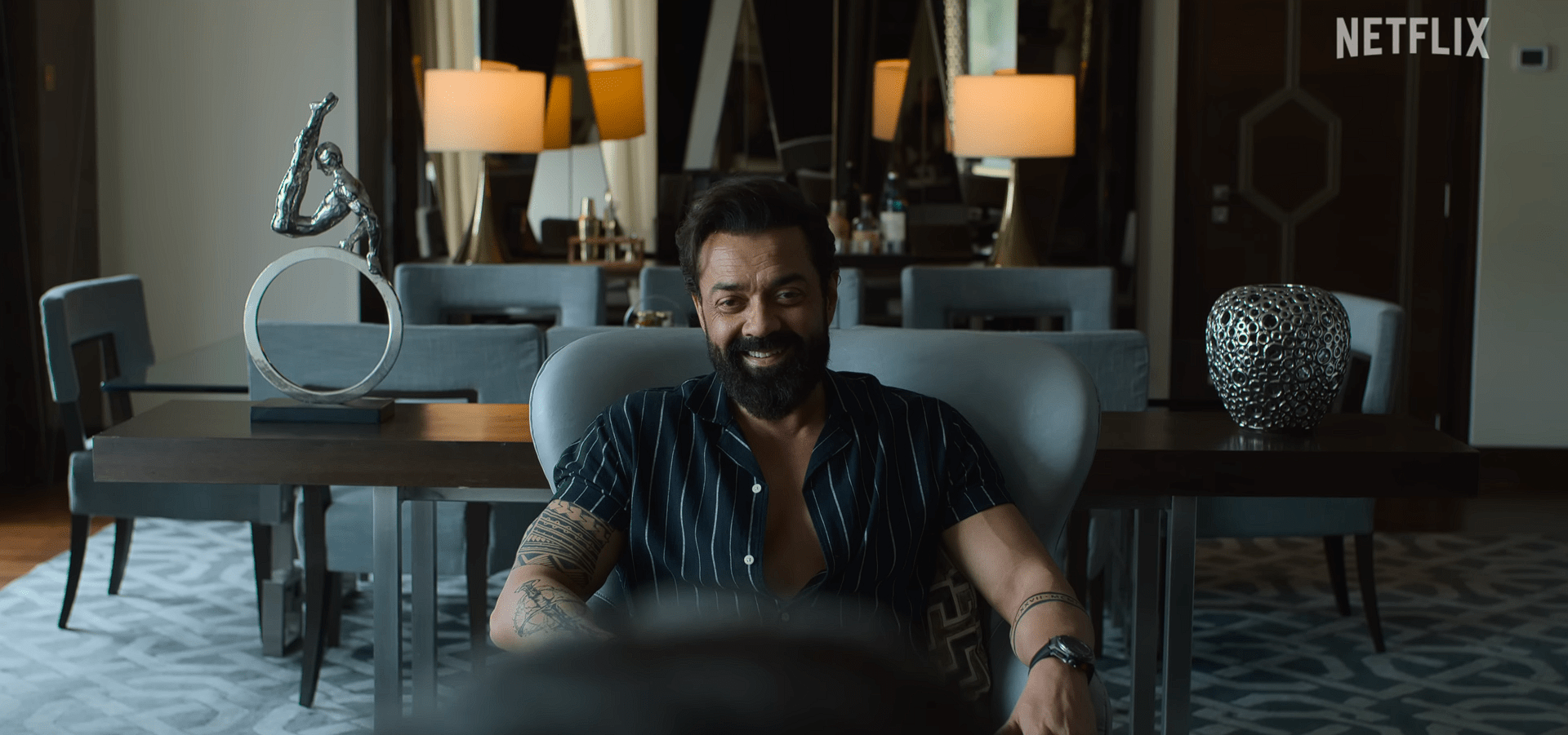 Bobby Deol man in a dark striped shirt smiles while sitting in a luxurious living room with modern décor and warm lighting.