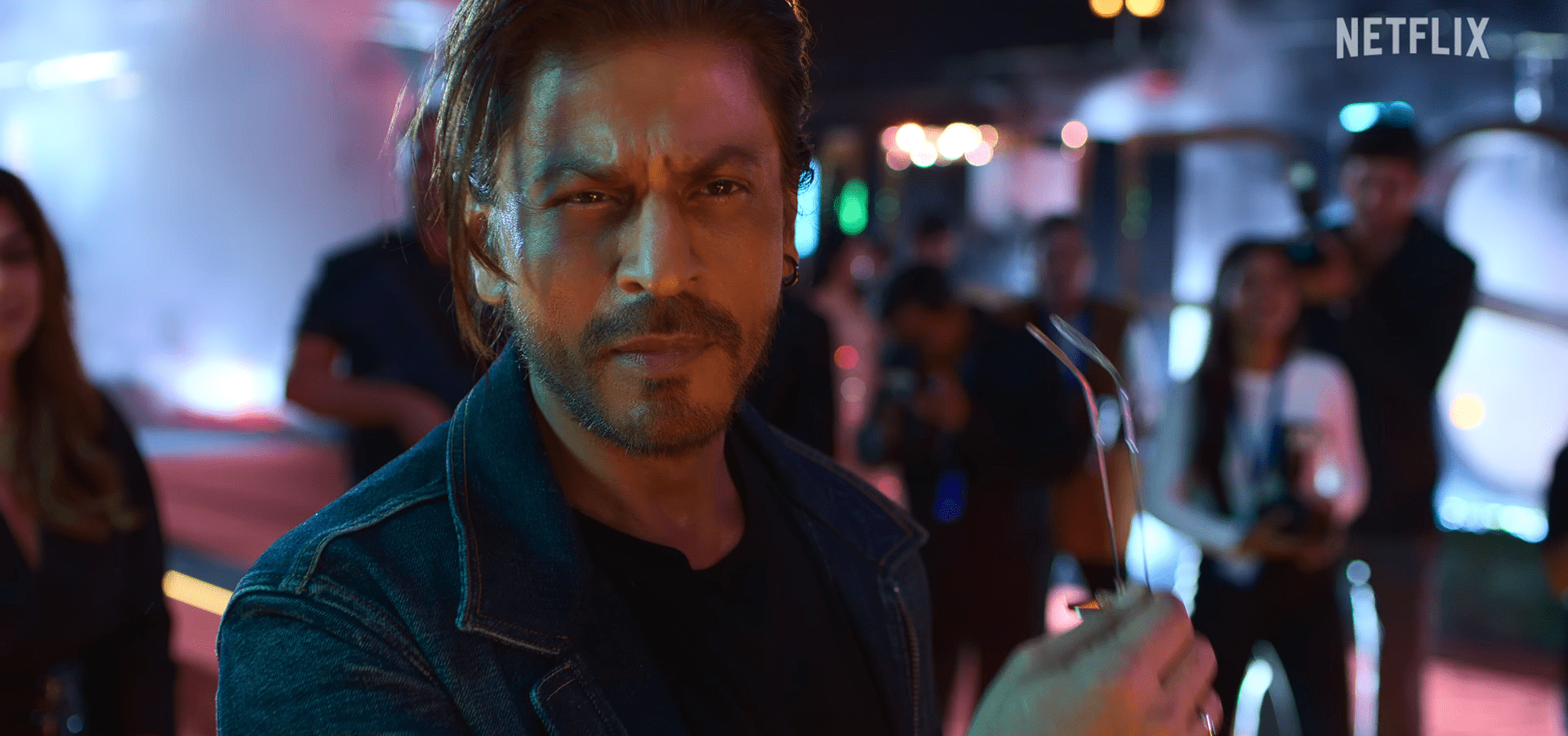 SRK in a denim jacket looks intensely at the camera while holding a pair of sunglasses, with bright lights and a crowd blurred in the background.